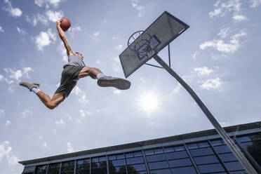 Young man dunking ball in hoop while playing basketball against sky on sunny day - STSF02621