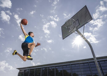 Junger männlicher Sportler, der beim Basketballspielen an einem sonnigen Tag den Ball in den Reifen eintaucht - STSF02622