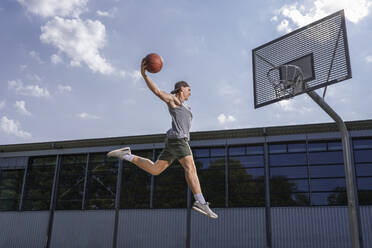 Junger männlicher Sportler, der an einem sonnigen Tag übt, den Ball in den Basketballkorb zu werfen - STSF02623