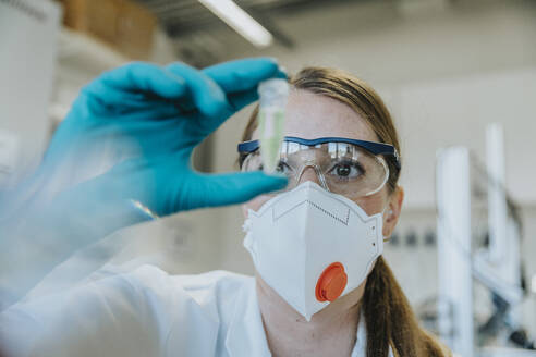 Young woman wearing protective face mask and eyeglasses examining test tube at laboratory - MFF06451