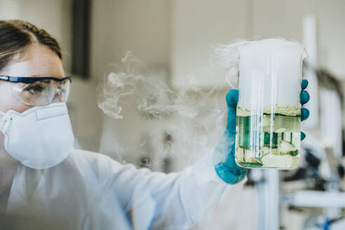 Laborant mit Schutzmaske und Brille, der einen Becher mit Flüssigkeit hält, während er im Labor steht - MFF06454