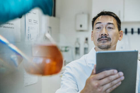 Wissenschaftler, der einen Flüssigkeitskolben betrachtet, während er ein digitales Tablet im Labor benutzt - MFF06480