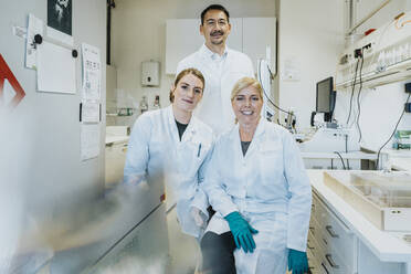 Coworkers smiling while sitting with scientist standing in background at laboratory - MFF06485