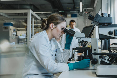 Young woman analyzing human brain microscope slide under microscope while sitting with scientists in background at laboratory - MFF06494