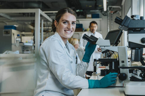 Smiling woman analyzing human brain slide under microscope while sitting with scientists in background at laboratory - MFF06495