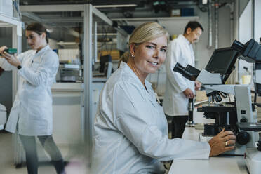 Smiling scientist sitting by microscope with coworkers working in background at laboratory - MFF06496