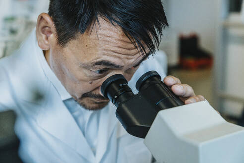 Male scientist looking though microscope while man standing in background at laboratory - MFF06499
