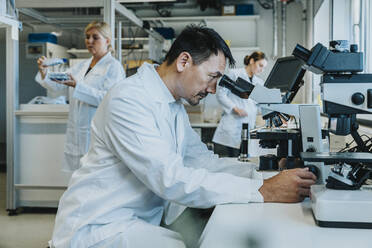 Male scientist sitting by microscope while coworker working in background at laboratory - MFF06501