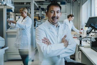 Male scientist sitting with arms crossed by microscope while coworker working in background at laboratory - MFF06503