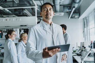 Wissenschaftlerin mit Hand in der Tasche und digitalem Tablet, stehend mit Mitarbeitern im Hintergrund im Labor - MFF06557