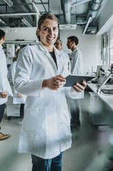 Lächelnde Frau, die ein digitales Tablet benutzt, während sie mit einem Mitarbeiter im Hintergrund in einem Labor steht - MFF06560