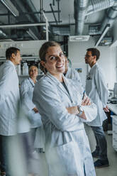Junge Wissenschaftlerin lächelt, während sie mit verschränkten Armen und einem Mitarbeiter im Hintergrund im Labor steht - MFF06563