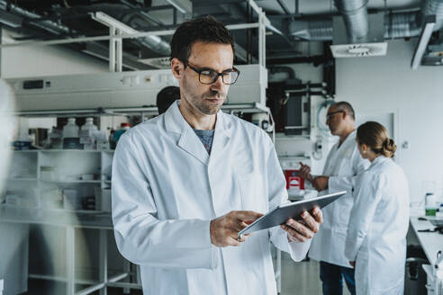 Scientist wearing eyeglasses using digital tablet while standing with coworker in background at laboratory - MFF06567