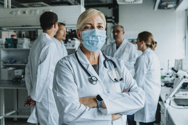 Selbstbewusster Wissenschaftler mit Gesichtsmaske, der mit verschränkten Armen und einem Mitarbeiter im Hintergrund im Labor steht - MFF06572