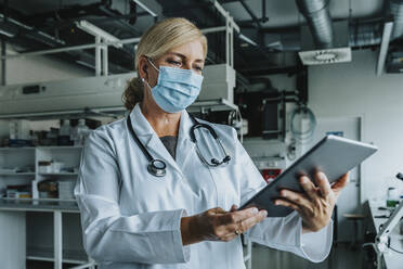 Scientist wearing face mask using digital tablet while standing at laboratory - MFF06576