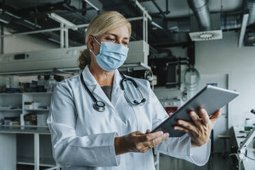 Wissenschaftlerin mit Gesichtsmaske, die im Labor stehend ein digitales Tablet benutzt - MFF06576