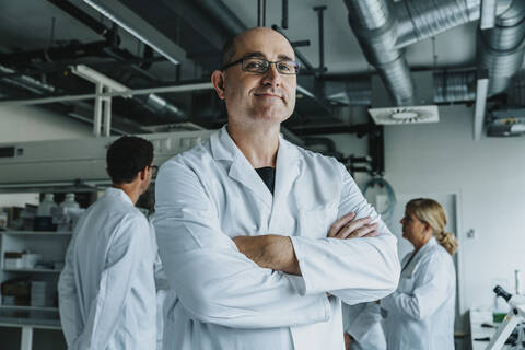 Selbstbewusster Wissenschaftler mit einem Mitarbeiter im Hintergrund im Labor stehend, lizenzfreies Stockfoto