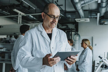 Wissenschaftler arbeitet an einem digitalen Tablet, während er mit einem Kollegen im Hintergrund im Labor steht - MFF06582