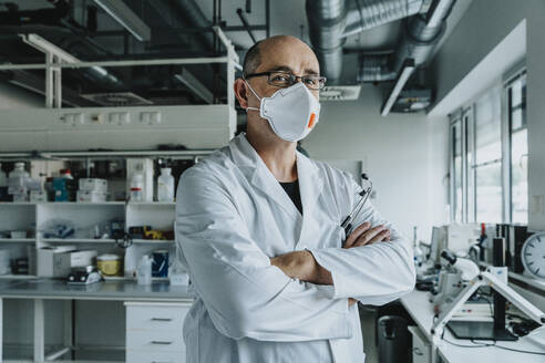 Confident scientist wearing face mask standing with arms crossed at laboratory - MFF06584