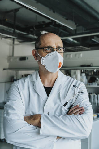 Scientist wearing face mask looking away while standing with arms crossed at laboratory stock photo