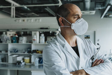 Male scientist wearing face mask looking away while standing with arms crossed at laboratory - MFF06586