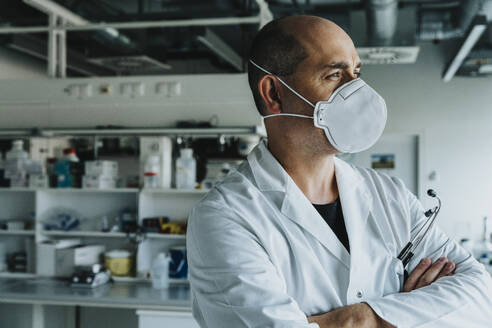 Männlicher Wissenschaftler mit Gesichtsmaske, der mit verschränkten Armen im Labor steht und wegschaut - MFF06586