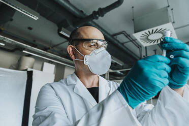 Wissenschaftler mit Schutzmaske und Brille, der einen Impfstoff in der Hand hält, während er im Labor steht - MFF06587