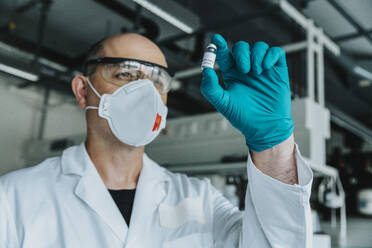 Wissenschaftler mit Schutzmaske und Brille, der eine Impfstoffflasche hält, während er im Labor steht - MFF06589