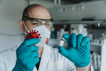 Scientist with face mask holding vaccine and coronavirus model while standing at laboratory - MFF06590