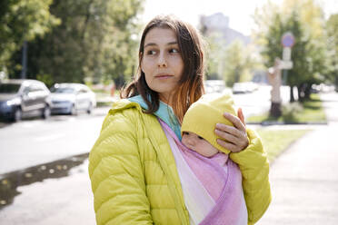 Mutter hält ihr in eine Decke eingewickeltes Baby, während sie an einem sonnigen Tag auf der Straße steht - EYAF01369