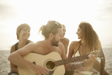 Mittlerer Erwachsener, der Gitarre spielt, während seine Freunde sitzend im Sand am Strand singen - AJOF00361