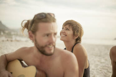Lächelnder Mann spielt Gitarre am Strand sitzend an einem sonnigen Tag - AJOF00362