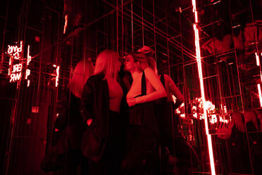 Female friends kissing while standing in red light room - DAMF00606