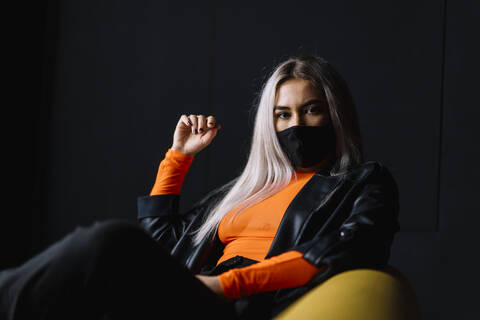 Young woman wearing protective face mask sitting on sofa against black wall stock photo