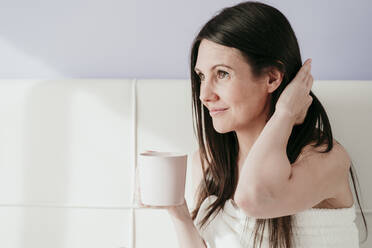 Thoughtful woman holding coffee cup while sitting in bedroom at home - EBBF00998