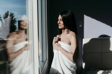 Smiling thoughtful woman wrapped in towel standing by window at home - EBBF01003
