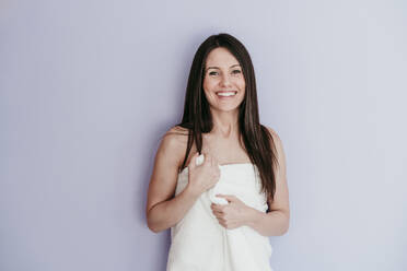 Smiling beautiful woman wrapped in towel standing against wall at home - EBBF01004