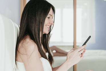 Lächelnde Frau mit Smartphone im Schlafzimmer zu Hause - EBBF01012