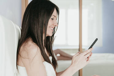 Smiling woman using smart phone in bedroom at home - EBBF01012