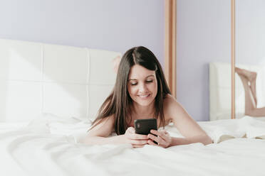 Smiling beautiful woman text messaging while lying on bed at home - EBBF01016