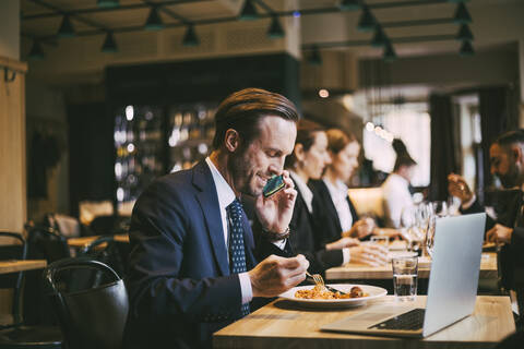 Smiling male business person talking on phone while eating food in restaurant stock photo