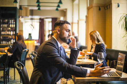 Side view of businessman talking on phone while using laptop in restaurant stock photo