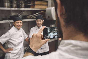 Male chef taking photograph of female coworkers in commercial kitchen - MASF20125