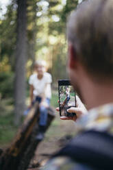 Father photographing daughter through smart phone in forest - MASF20186