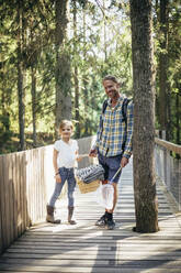 Portrait of father and daughter with picnic basket standing on footbridge in forest - MASF20197