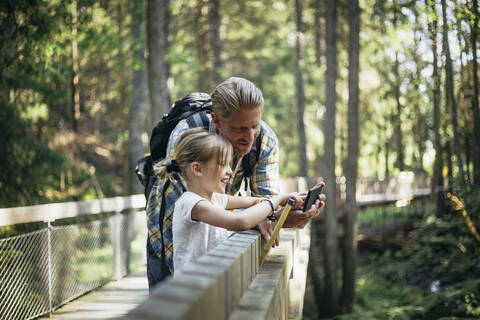 Lächelnder Vater mit Rucksack und Tochter, die im Wald auf ihr Smartphone schaut, lizenzfreies Stockfoto