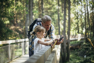 Smiling father with backpack and daughter looking at smart phone in forest - MASF20200