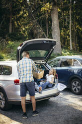 Rear view of father standing with picnic basket while daughter sitting in car trunk - MASF20207