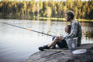 Portrait of smiling daughter fishing with father while sitting by lake - MASF20219