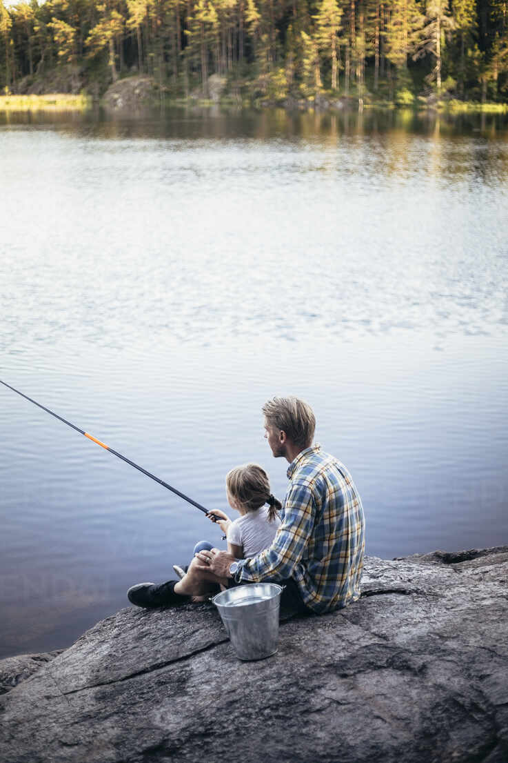Father and daughter fishing while sitting by lake stock photo
